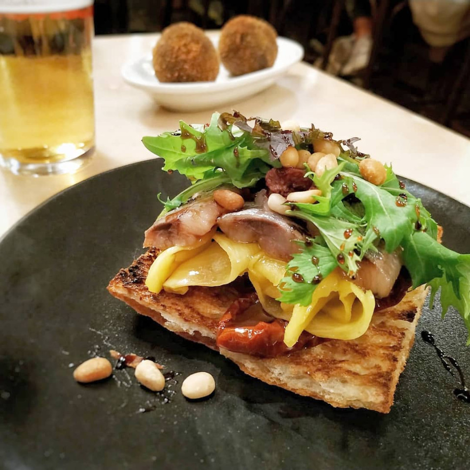 Bright, colourful tapa on black plate with beer and croquettes in background.